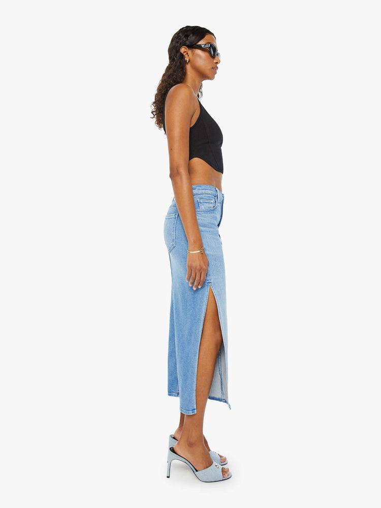 Side view of a woman in a light blue high-rise denim midi skirt with a diagonal thigh-high slit and a narrow fit with whiskering and fading at the knees. Styled with a black tank top.