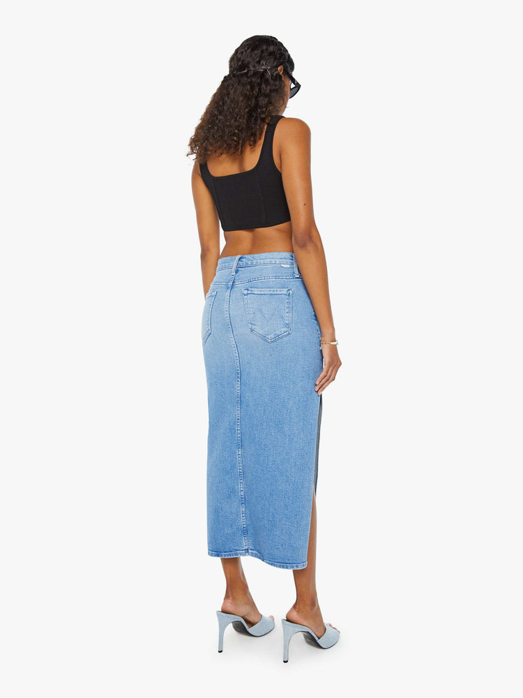Back view of a woman in a light blue high-rise denim midi skirt with a diagonal thigh-high slit and a narrow fit with whiskering and fading at the knees. Styled with a black tank top.