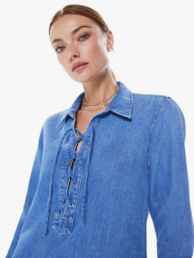 Close up view of a women collared shirt dress with a deep V-neck that laces and ties, long sleeves and a playfully short curved hem in a mid-blue wash.