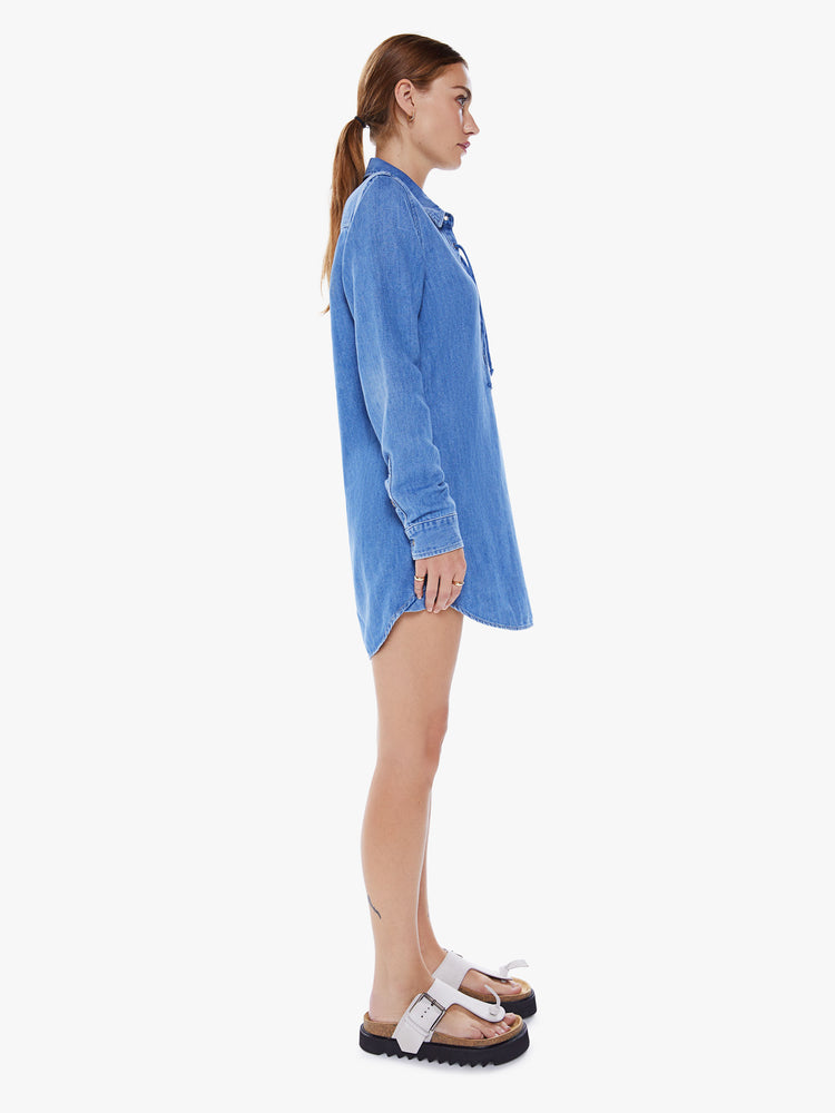Side view of a women collared shirt dress with a deep V-neck that laces and ties, long sleeves and a playfully short curved hem in a mid-blue wash.