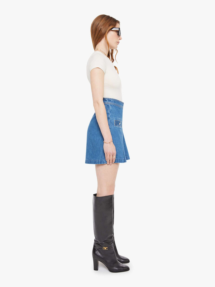 Side view of a woman in a med blue  denim mini skirt with a high rise, slit pockets, wide pleats and a zip closure in the back. 