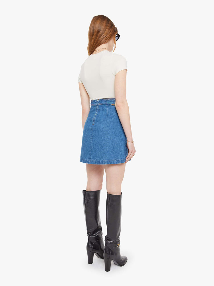 Back view of a woman in a med blue  denim mini skirt with a high rise, slit pockets, wide pleats and a zip closure in the back. 