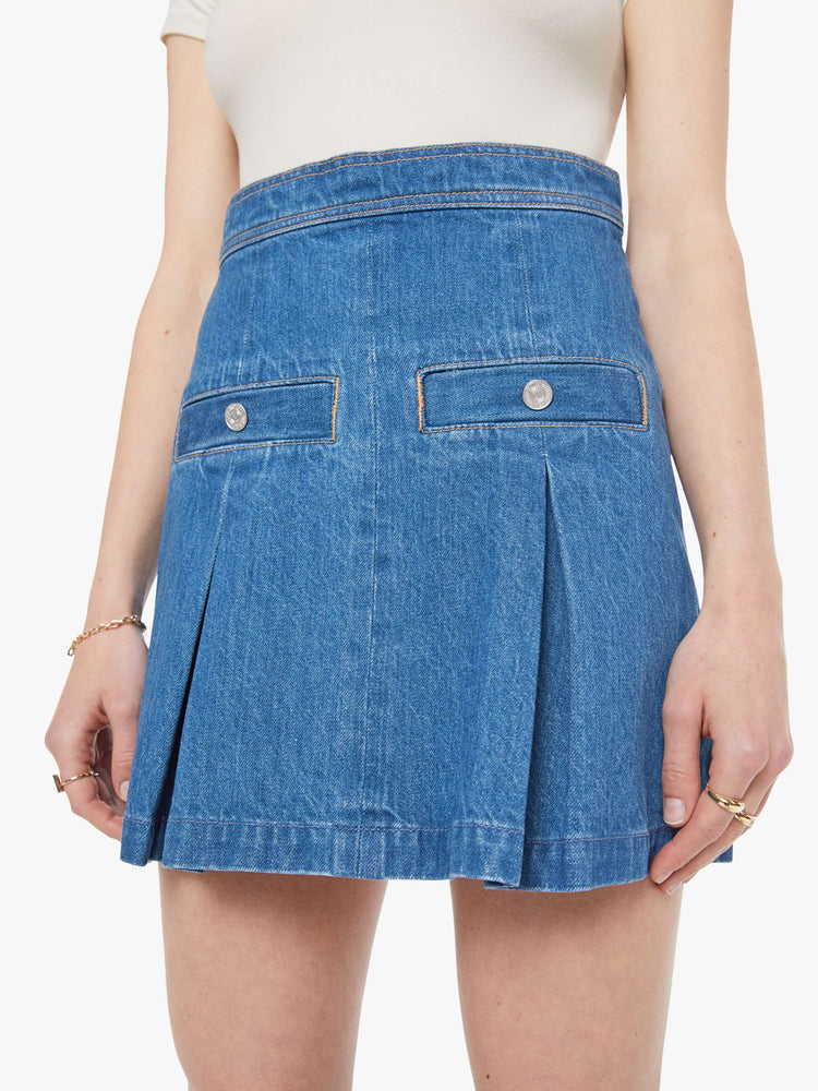 Close up view of a woman in a med blue  denim mini skirt with a high rise, slit pockets, wide pleats and a zip closure in the back. 