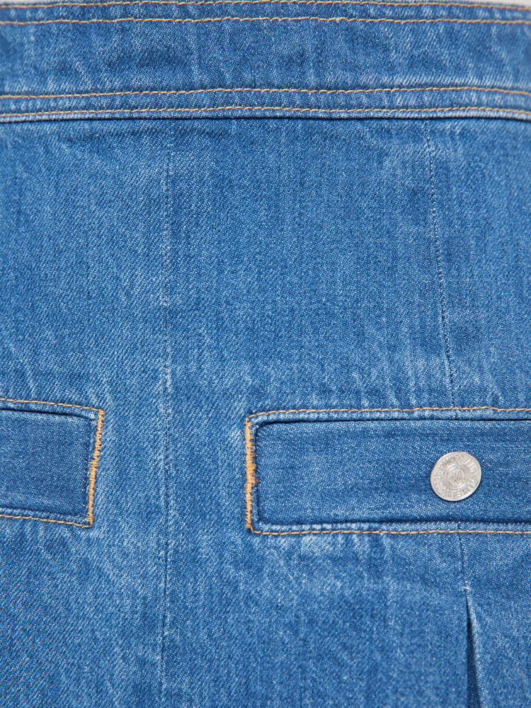 Swatch view of a woman in a med blue  denim mini skirt with a high rise, slit pockets, wide pleats and a zip closure in the back. 