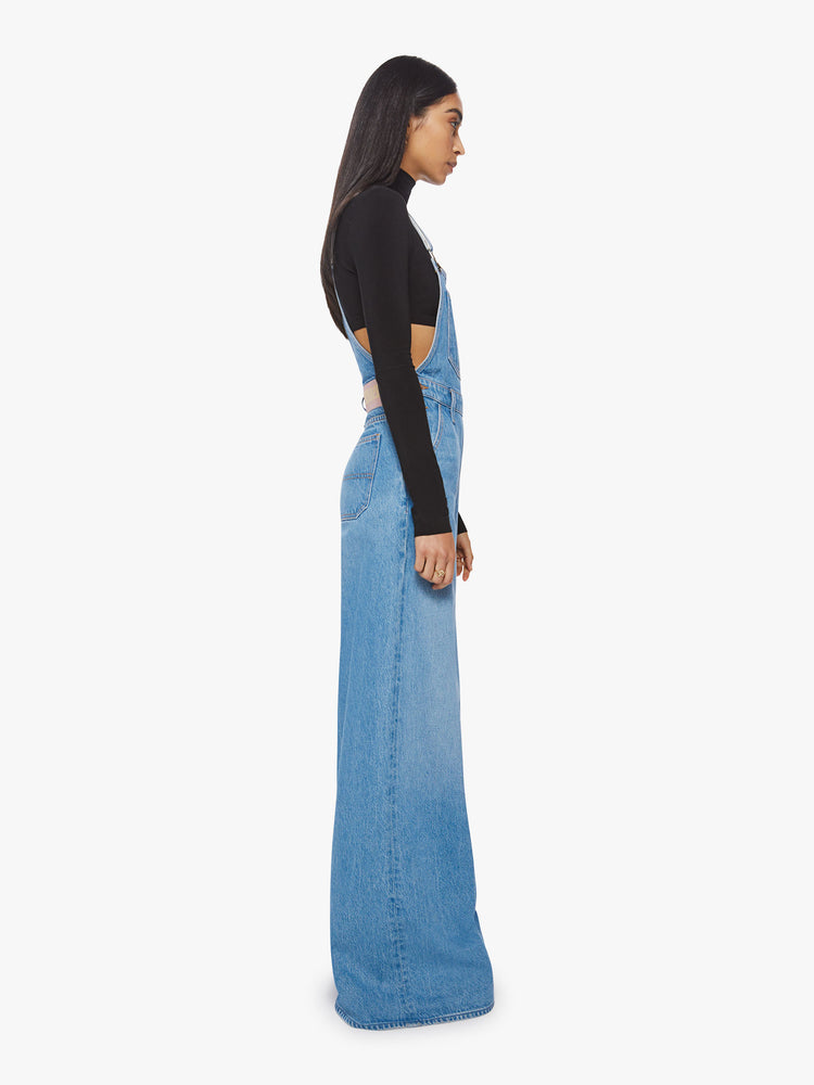 Side view of a woman mid-blue wide leg overalls feature a loose, full leg, a hammer loop and a 32-inch inseam.