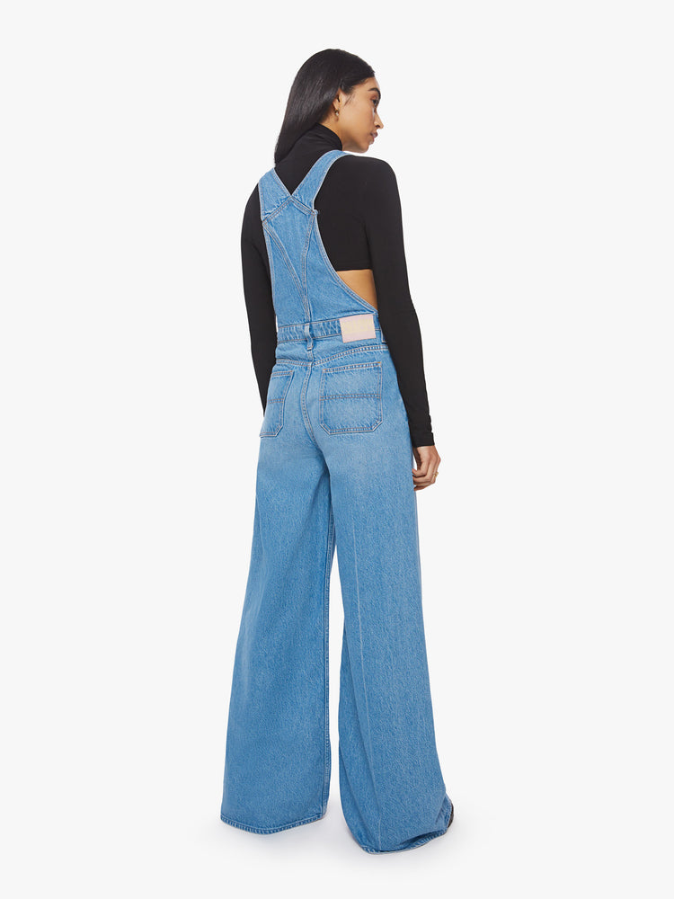 Back view of a woman mid-blue wide leg overalls feature a loose, full leg, a hammer loop and a 32-inch inseam.