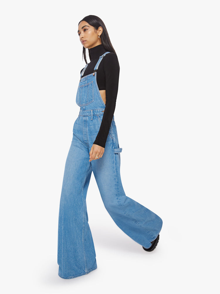 Walking view of a woman mid-blue wide leg overalls feature a loose, full leg, a hammer loop and a 32-inch inseam.