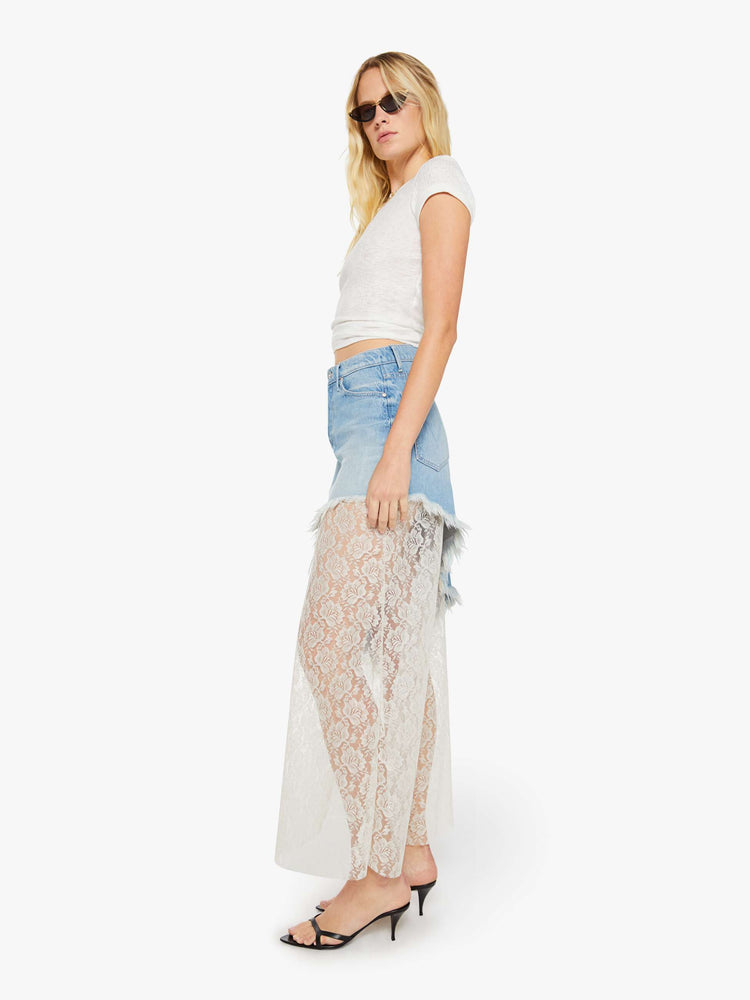 Right side view of a woman in a light blue denim maxi skirt with a high rise, button fly, angled frayed hem and sheer lace slip underneath that hits at the ankles. 