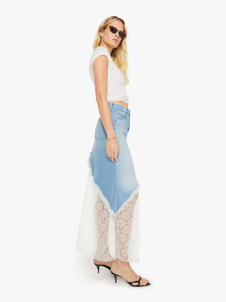 Side view of a woman in a light blue denim maxi skirt with a high rise, button fly, angled frayed hem and sheer lace slip underneath that hits at the ankles. 