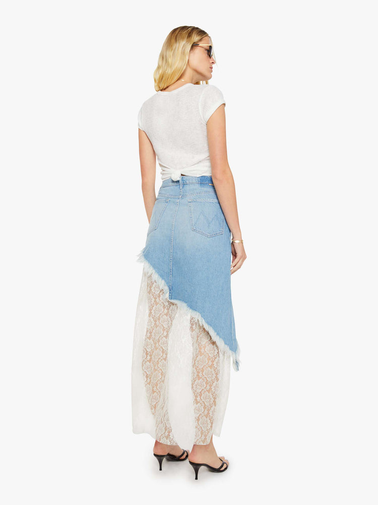 Back view of a woman in a light blue denim maxi skirt with a high rise, button fly, angled frayed hem and sheer lace slip underneath that hits at the ankles. 