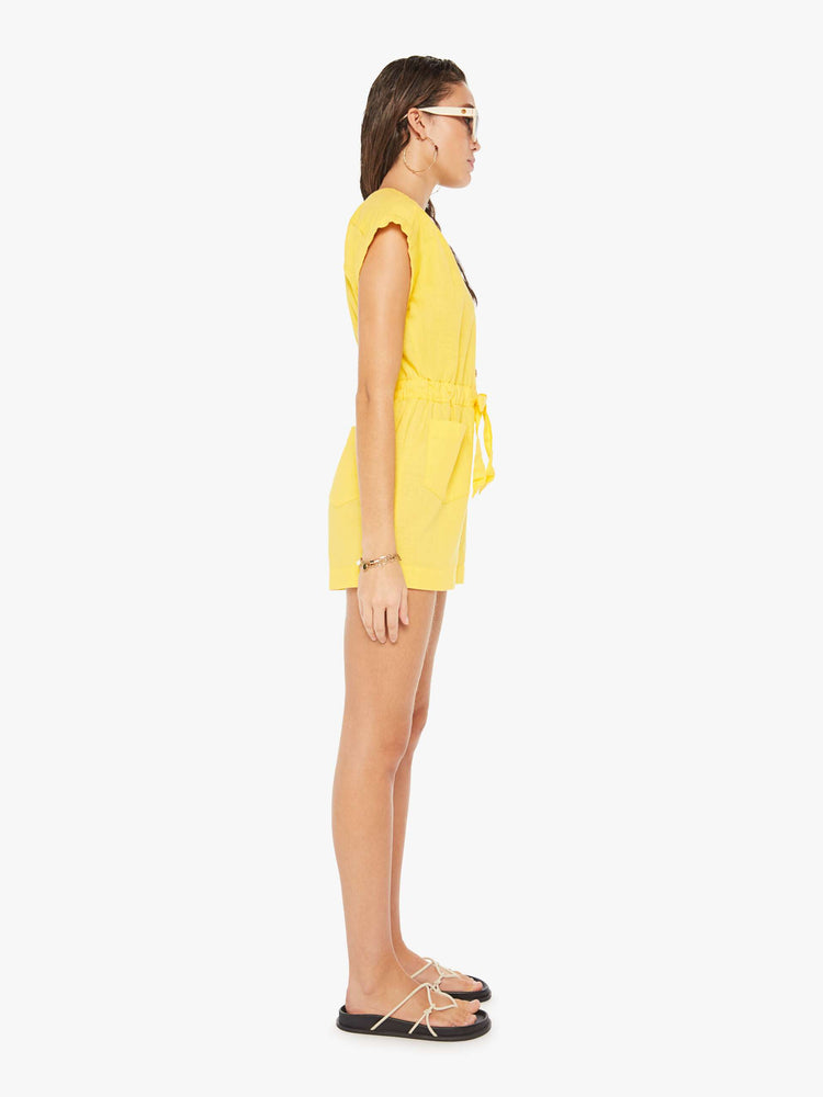 Side view of a woman wearing a short yellow romper featuring cuffed cap sleeves, a v neckline, and an elastic tie waist.