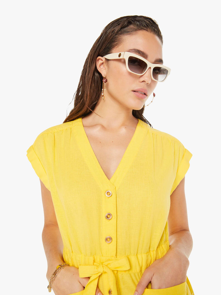 Front close up view of a woman wearing a short yellow romper featuring cuffed cap sleeves, a v neckline, and an elastic tie waist.