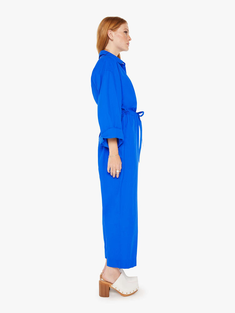 Side view of a woman royal blue lightweight collared jumpsuit with longsleeves, a tied waist, cargo patch pockets and an ankle-length straight leg.