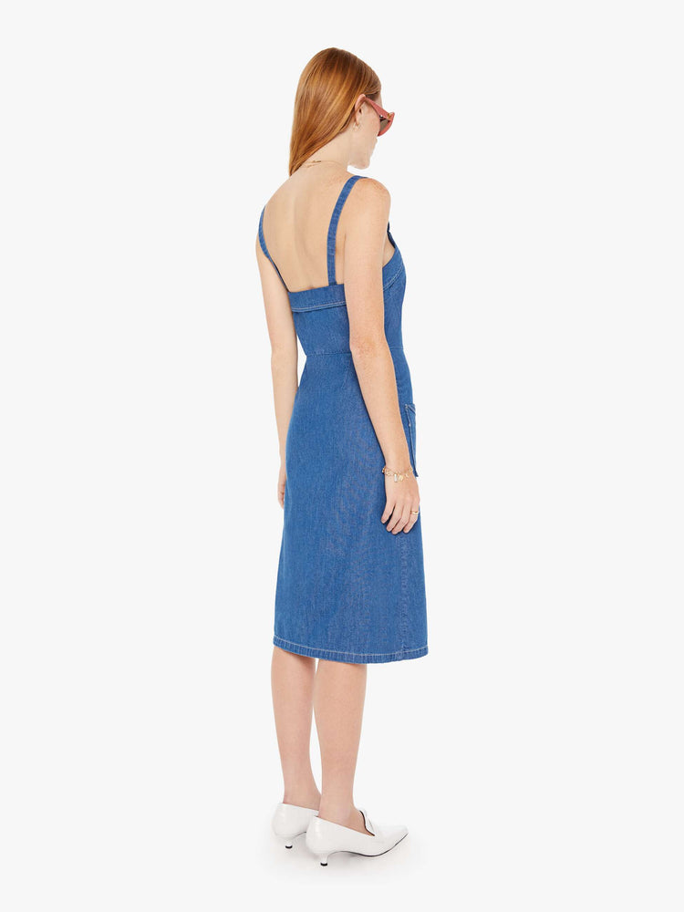 Back view of a woman in a mid blue denim midi dress with a V-neck, thin straps and folded details at the neck and pockets