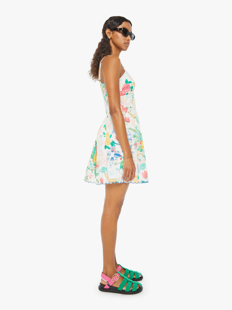 Side view of a woman in a sleeveless mini dress with a V-neck, fitted bodice, slit pockets, scalloped edges, and finished with hand-drawn doodles throughout and blue stitching at the hem.