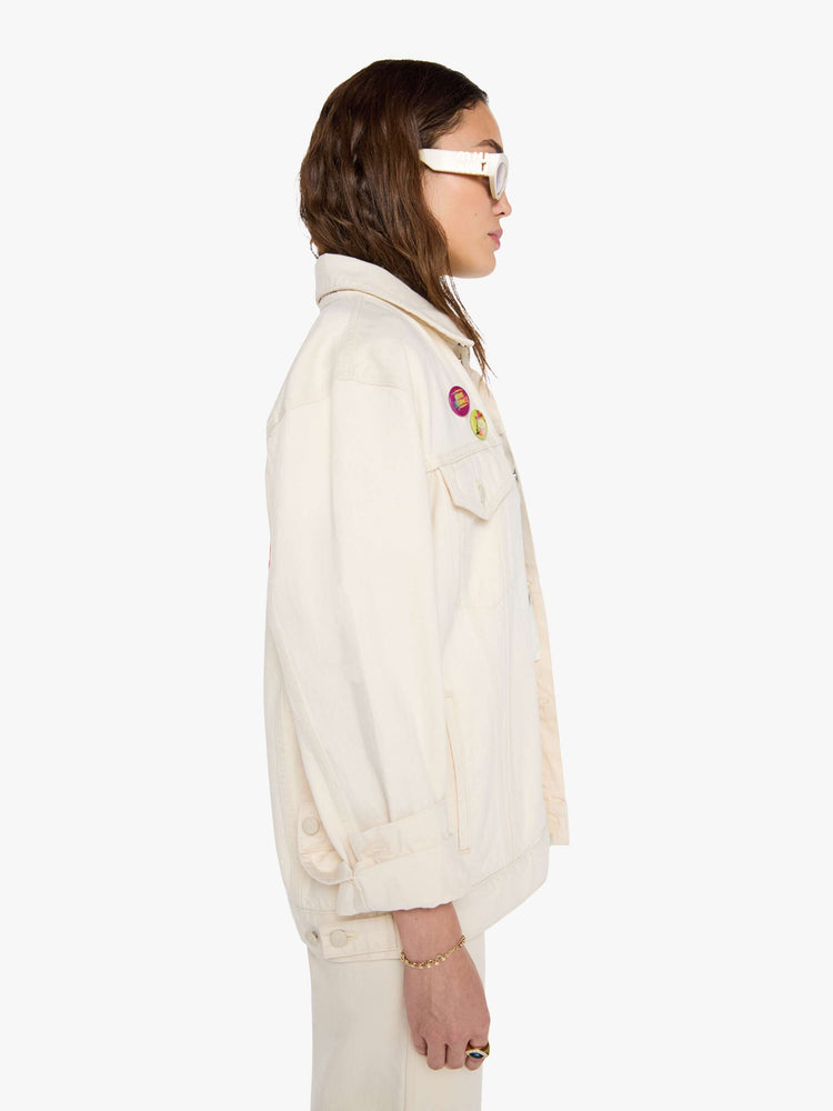 WOMAN side view in a  creamy white with tonal hardware, patches and pins denim jacket with drop shoulders, front patch pockets and a boxy fit.