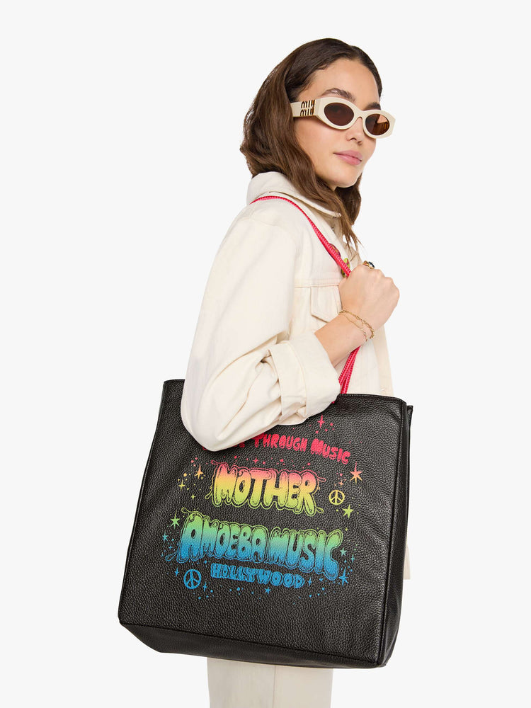 Woman holding a black faux leather with hot pink straps, the tote features a colorful Amoeba Music graphic and MOTHER's name in rainbow font.