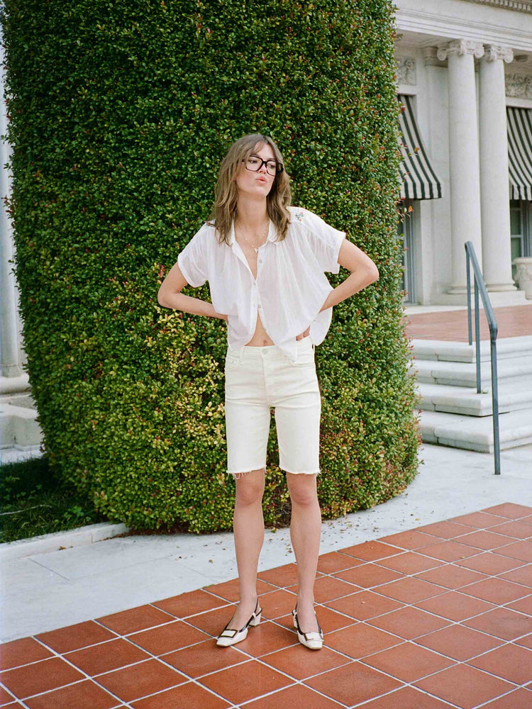 An editorial image of a woman standing in front of a large bush, wearing cream bermuda cut-off shorts and a white button down blouse.