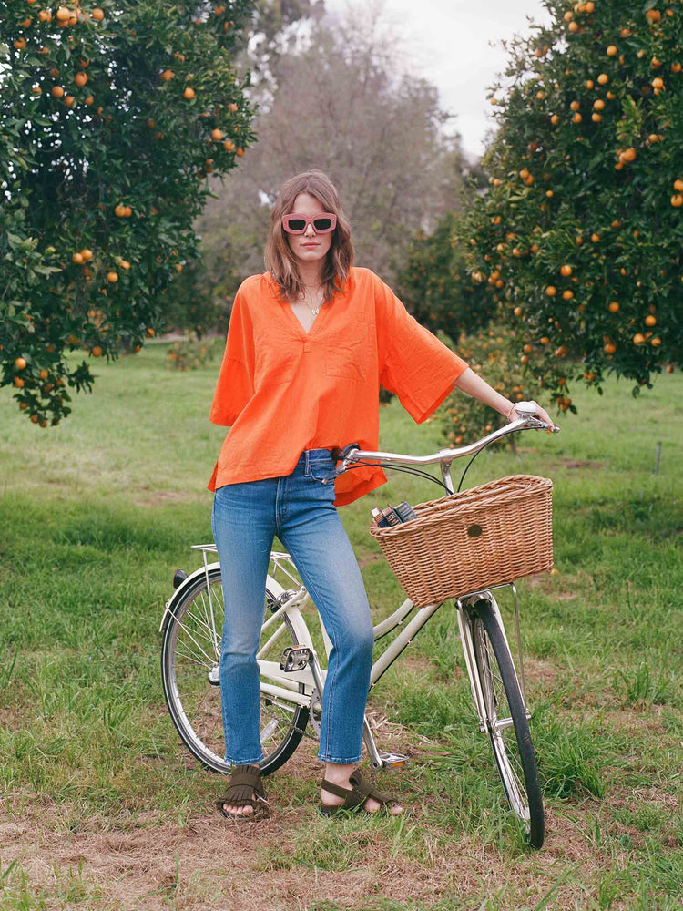 An editorial image of a woman standing next to a bicycle in an orange grove, wearing medium blue wash jeans and a bright orange top.