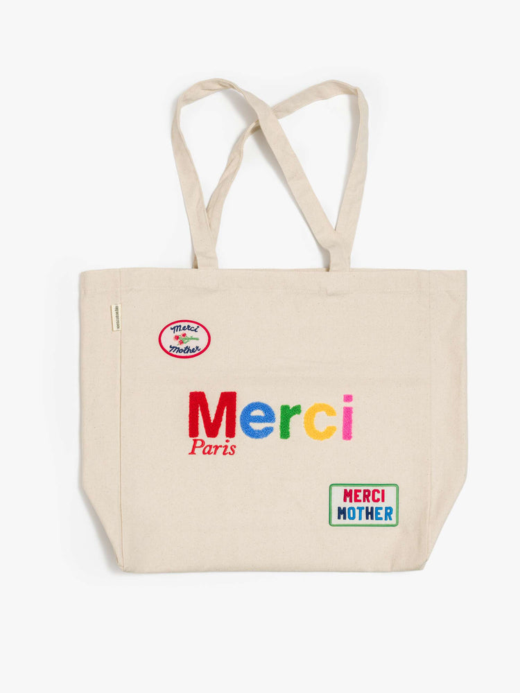 Flat view a cream tote with fuzzy multi-colored text and colorful patches at the bottom right and top left corners.