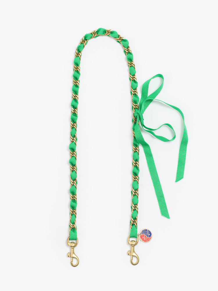 Flat view of a brass chain shoulder strap is detailed with a green ribbon and a small charm.