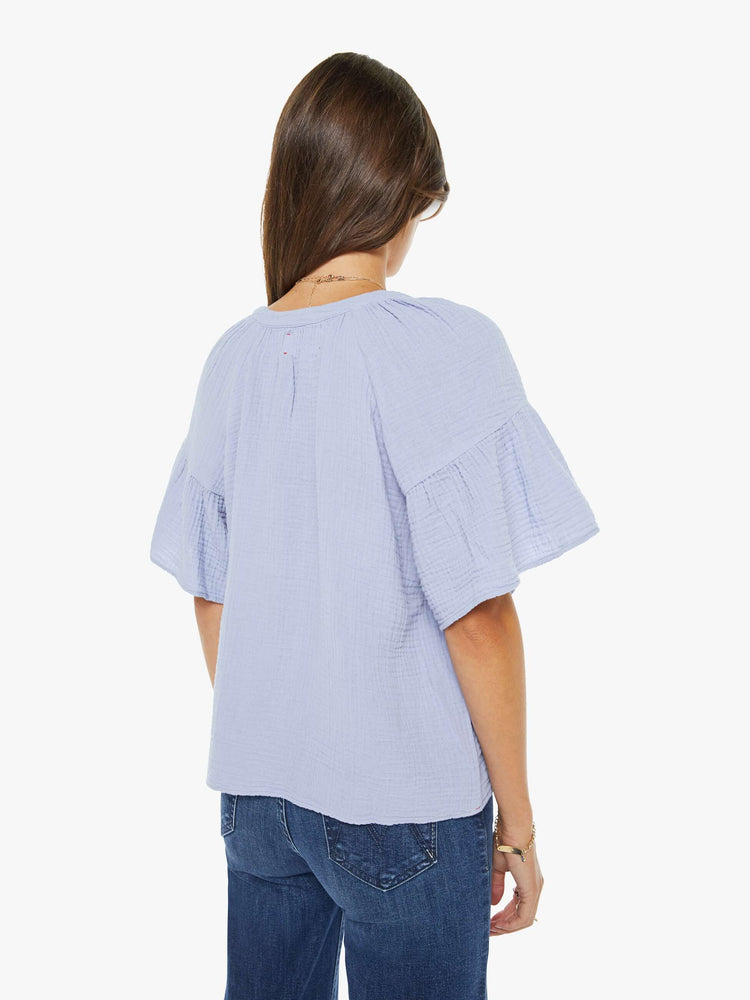 Back view of a woman in a light lavender with a deep V-neck, ruffled short sleeves and a loose, flowy fit.
