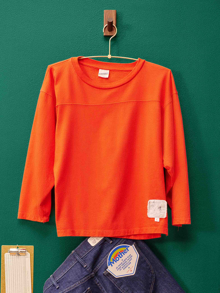 Lost & Found editorial image of a orange 3/4 sleeve tee hanging against a green wall.