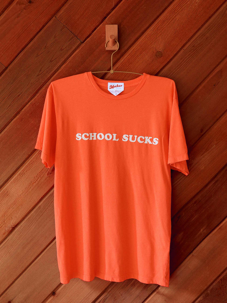 Lost & Found editorial image of an orange crew neck tee on a hanger with the graphic "SCHOOL SUCKS" against a wood panel wall.
