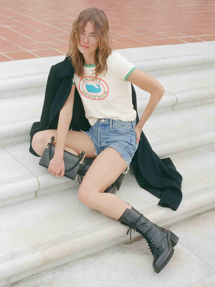 An editorial image of a woman sitting on a set of stairs, wearing blue cut-off jean shorts and a white graphic ringer tee.