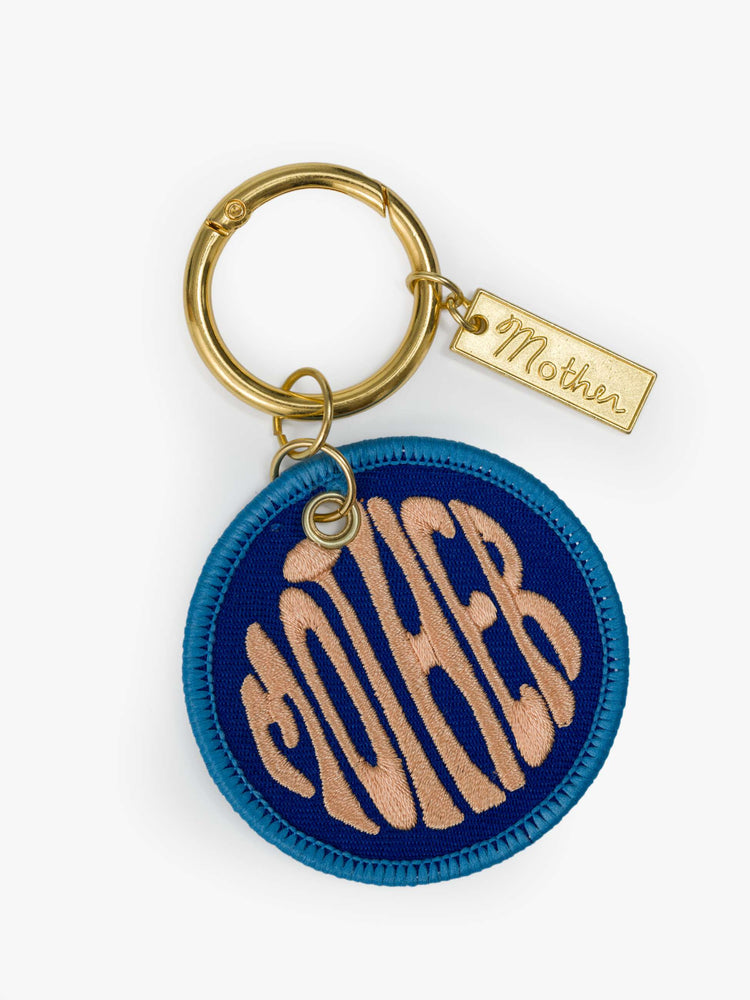 Front view of a gold-toned keychain with a circular M patch designed in blue and pink.