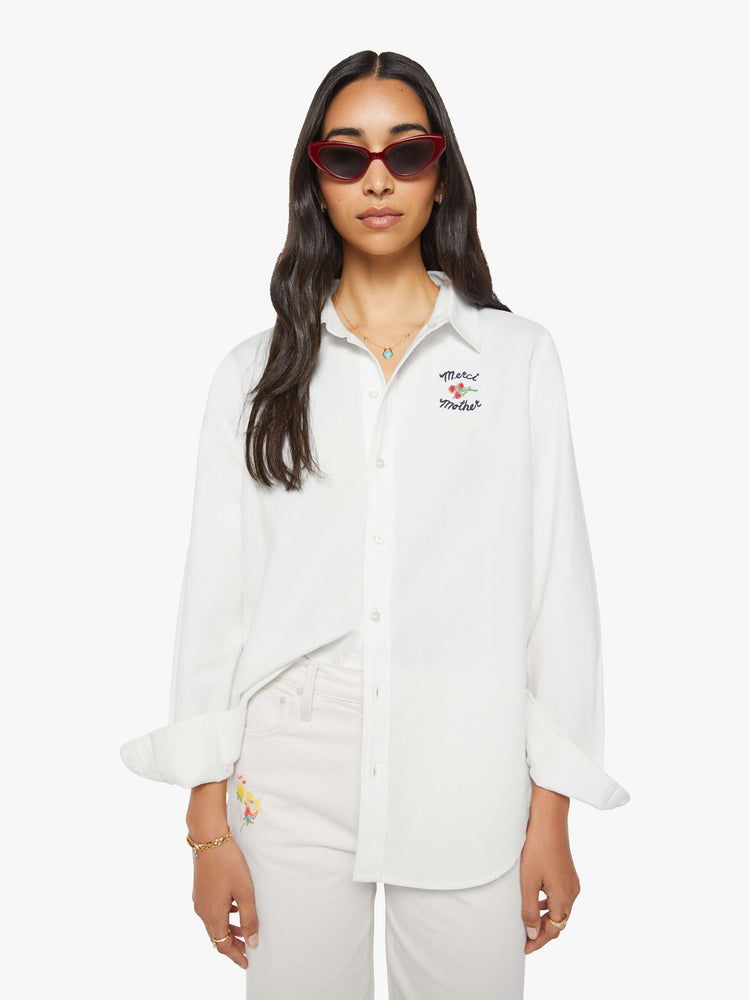 Front view of a woman in warm white button up with embroidered text in navy and a small bouquet on the chest.