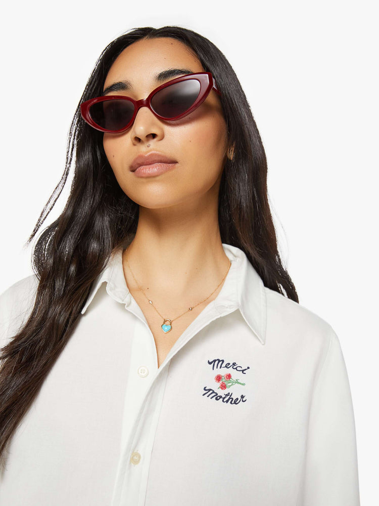 Close up view of a woman in warm white button up with embroidered text in navy and a small bouquet on the chest.