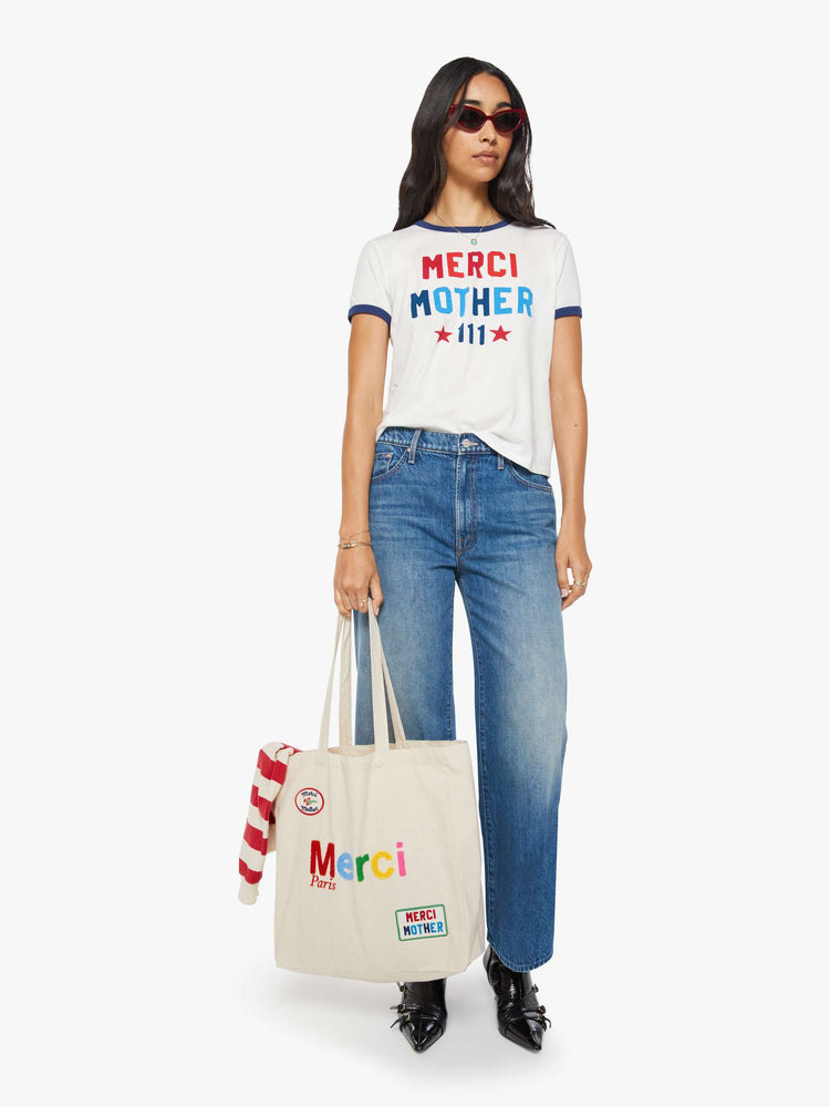 Full body view of a woman holding cream tote with fuzzy multi-colored text and colorful patches at the bottom right and top left corners.