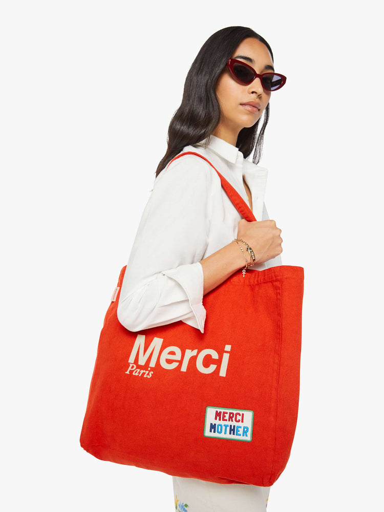 Side view of a woman holding view of a woman holding a red tote with bold flocked text in ecru, finished in a raised, velvety texture, and colorful patches at the bottom right and top left corners.