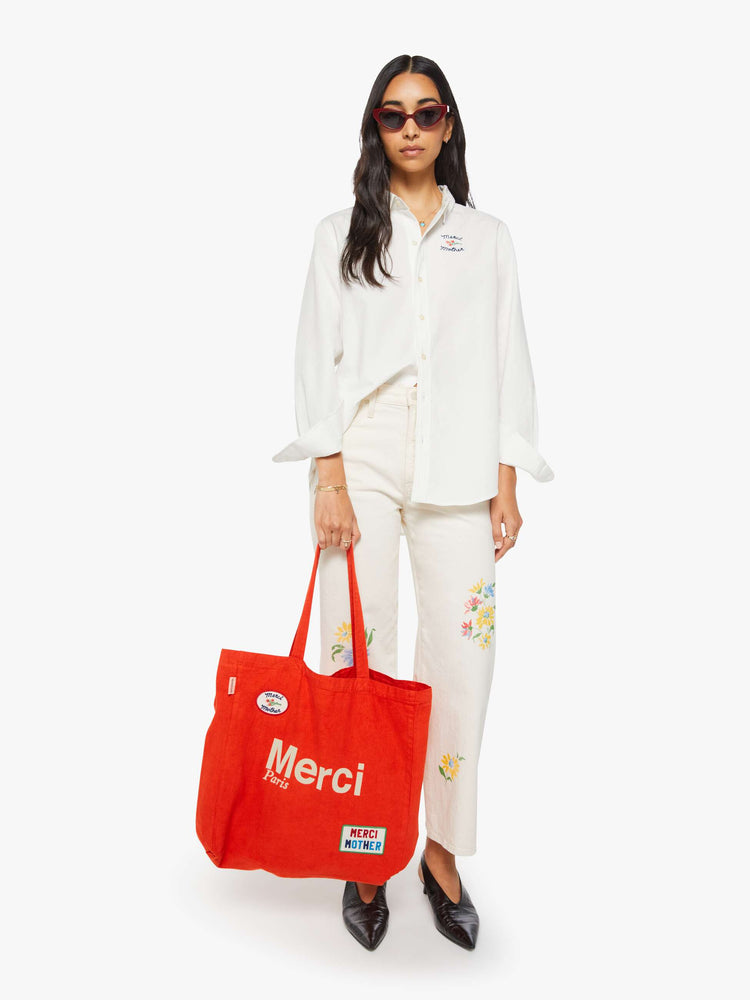 Full body view of a woman holding view of a woman holding a red tote with bold flocked text in ecru, finished in a raised, velvety texture, and colorful patches at the bottom right and top left corners.