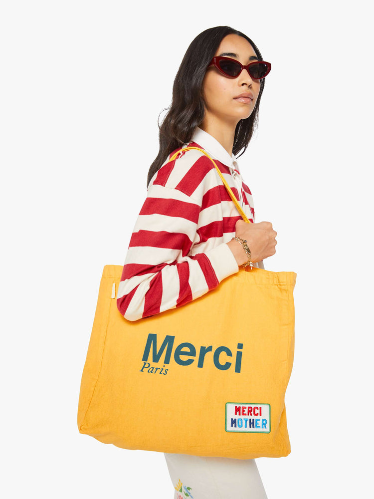 Side view of a woman holding a bright yellow tote with bold flocked text in blue, finished in a raised, velvety texture, and colorful patches at the bottom right and top left corners.