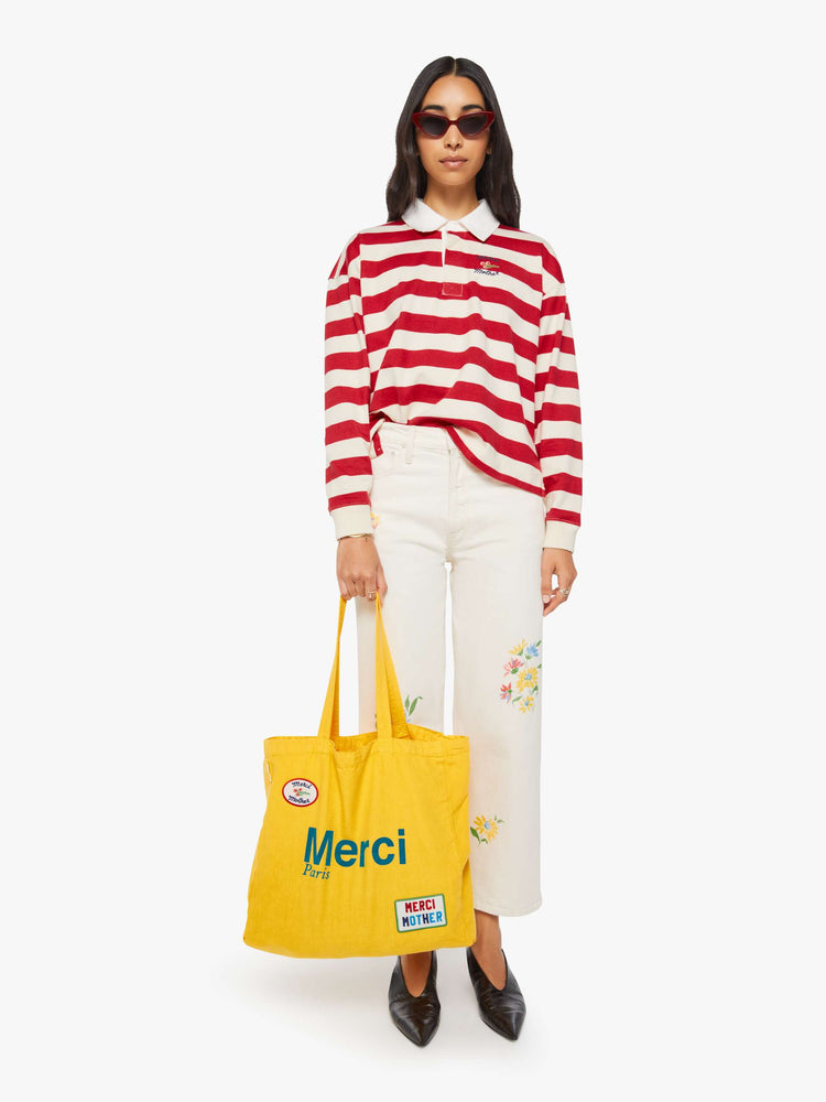 Full body view of a woman holding a bright yellow tote with bold flocked text in blue, finished in a raised, velvety texture, and colorful patches at the bottom right and top left corners.