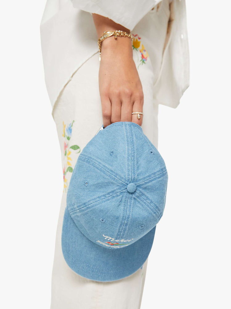 On model holding view of a light denim hat with embroidered text in white and a bouquet of red flowers.