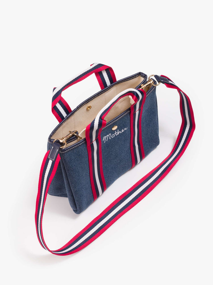 Flat view of a classic blue denim with red and white trim, top handles, an optional 34-inch shoulder strap, a snap closure and MOTHER embroidered across the front in white.