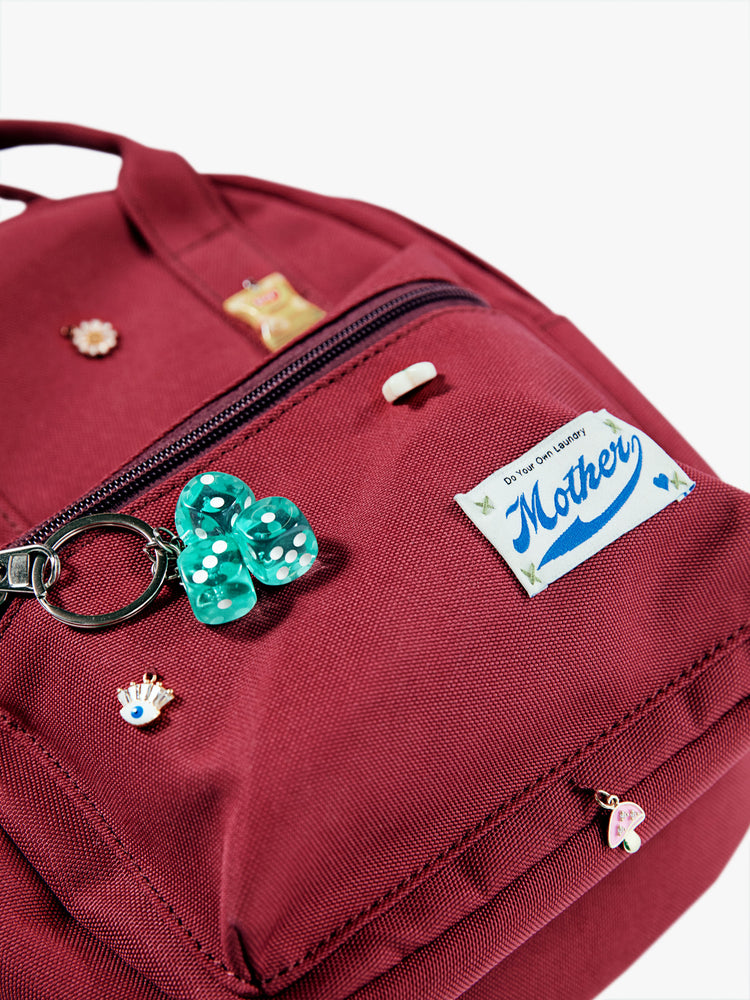 Flat image of a maroon mini backpack featuring assorted charms and a dice keychain.