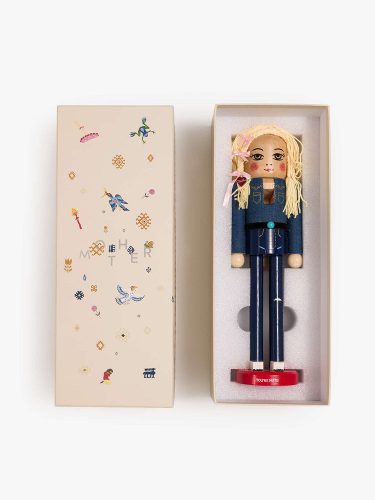 In box view of a traditional nutcracker a 70s-inspired twist with a Canadian tuxedo, turquoise-detailed belt, blonde braids, pink bows and a MOTHER heart charm.