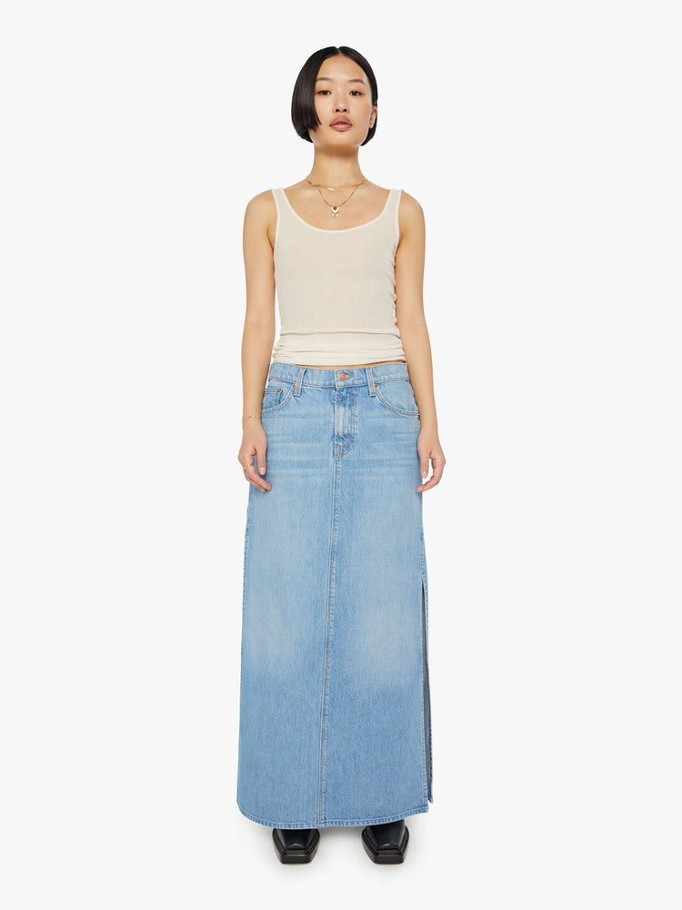 A front view of a woman wearing a medium blue wash denim maxi skirt featuring a mid rise and side slit, paired with a sheer tank top.