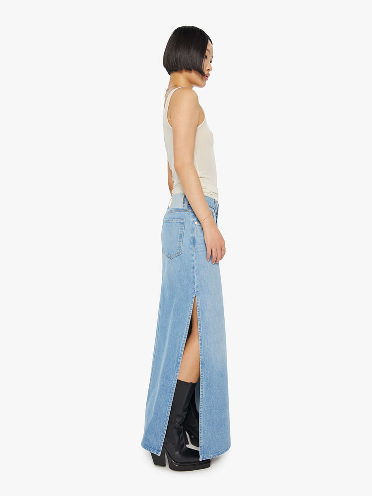 A side view of a woman wearing a medium blue wash denim maxi skirt featuring a mid rise and side slit, paired with a sheer tank top.