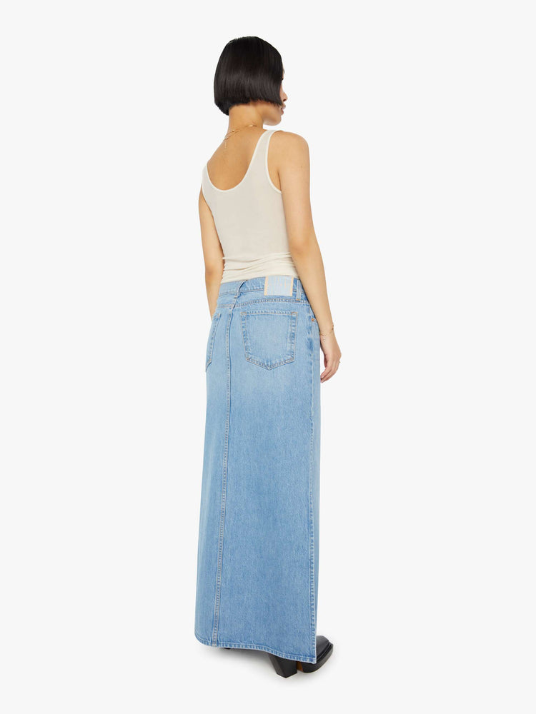 A back view of a woman wearing a medium blue wash denim maxi skirt featuring a mid rise and side slit, paired with a sheer tank top.