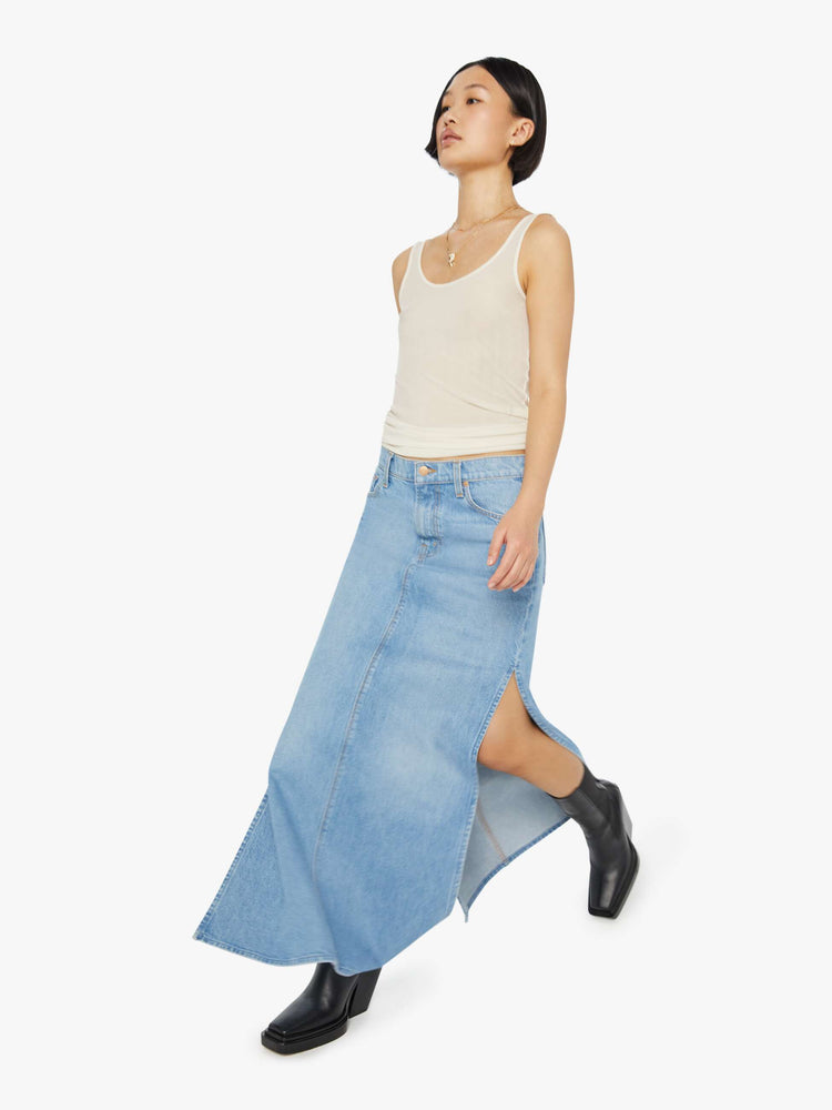 A front view of a woman walking in a medium blue wash denim maxi skirt featuring a mid rise and side slit, paired with a sheer tank top.