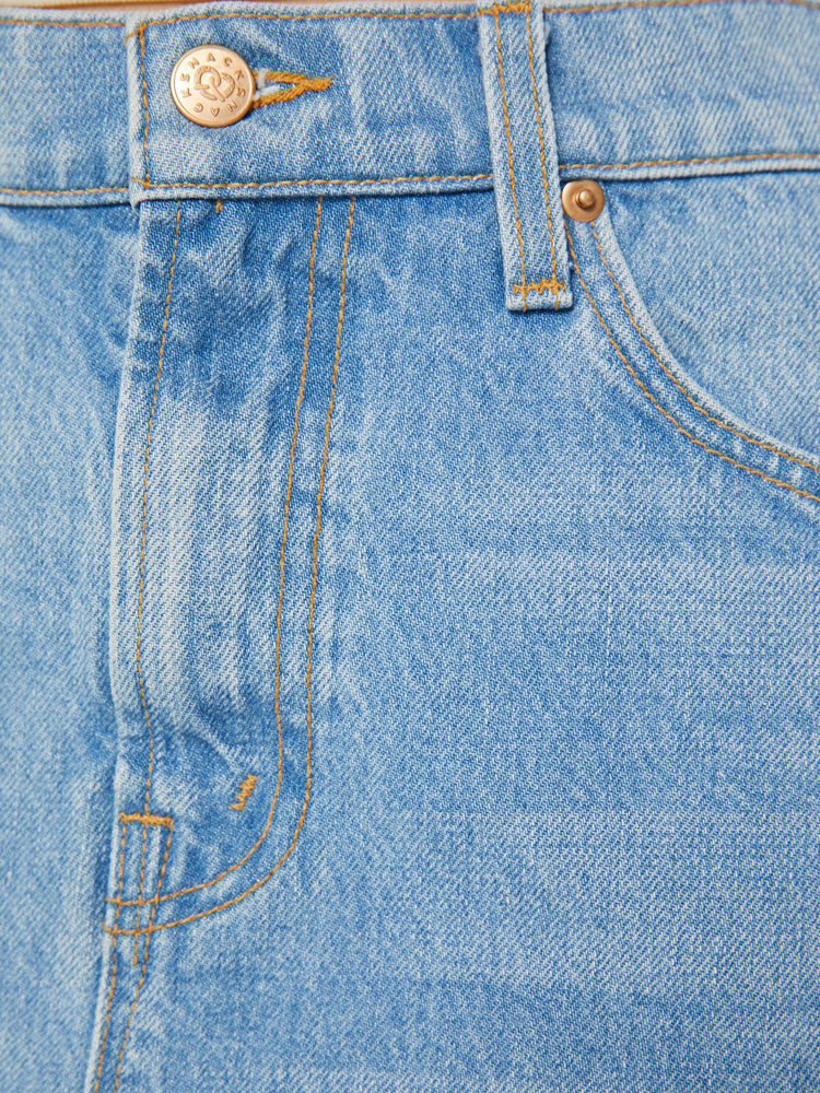 A close up swatch detail view of a medium blue wash denim featuring brass hardware and contrast brown thread.