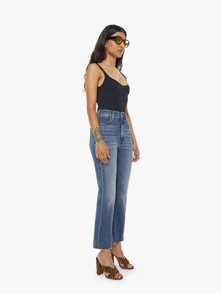 Side angle view of a petite woman mid blue wash jean with a high-rise flare has an ankle-length inseam and a raw hem.