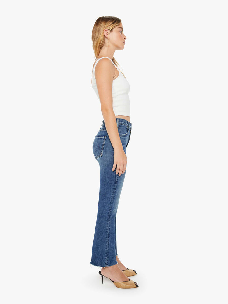 Side view of a petite woman mid blue wash jean with a high-rise flare has an ankle-length inseam and a raw hem.