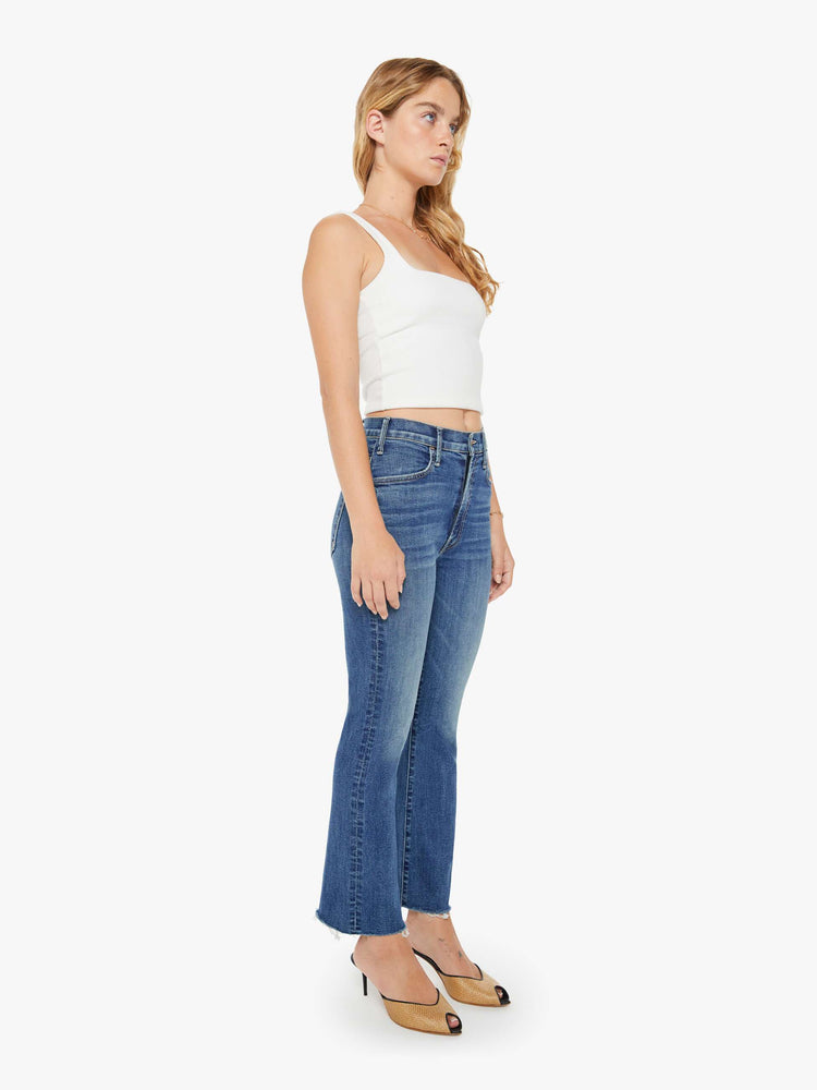 Side angle view of a petite woman mid blue wash jean with a high-rise flare has an ankle-length inseam and a raw hem.