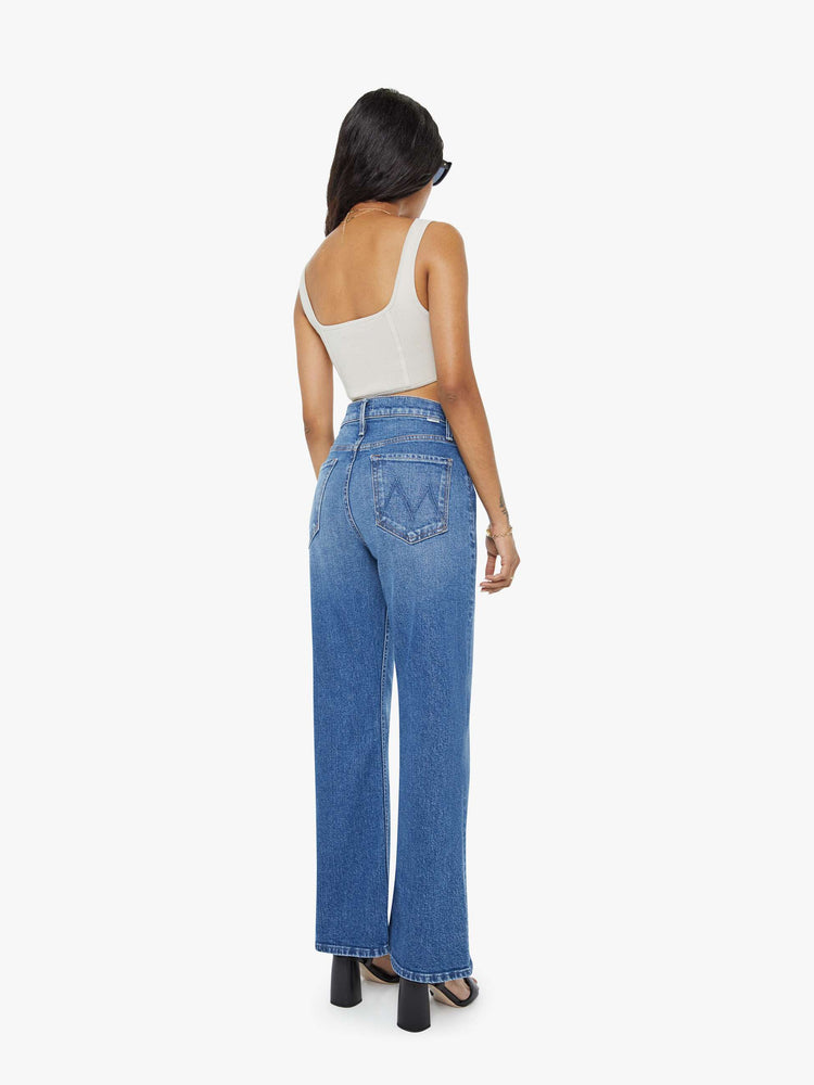 Back view of a petite woman in a med blue high-rise bootcut with a zip fly, looser leg and a long 29-inch inseam with a clean hem.